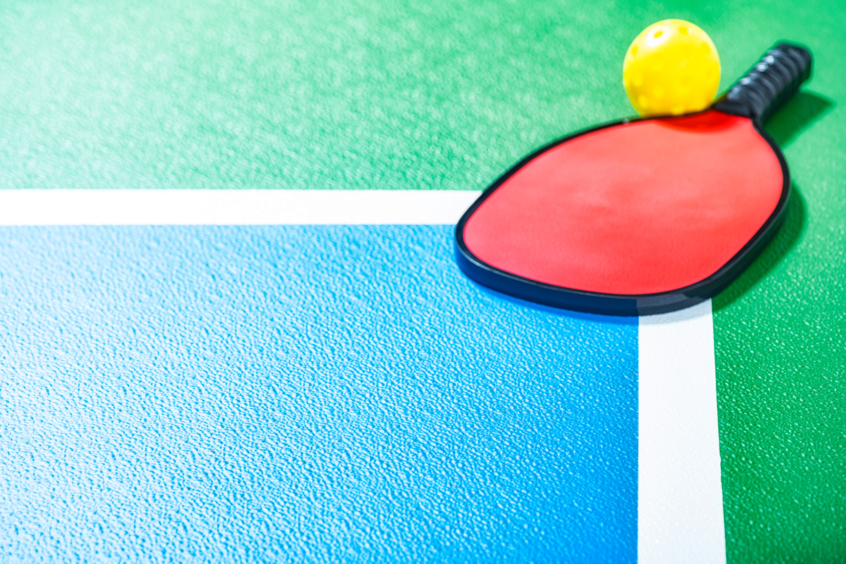 Selective focus on a Pickleball court with a red paddle and yellow ball in the corner